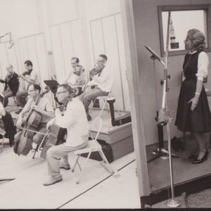 Patti Page sings along with the band in a recording studio booth.
